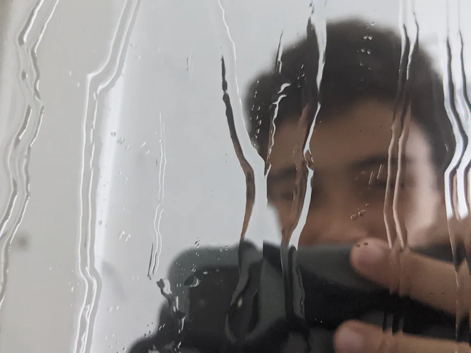 Reyaan behind rain-streaked glass, camera raised to his eye