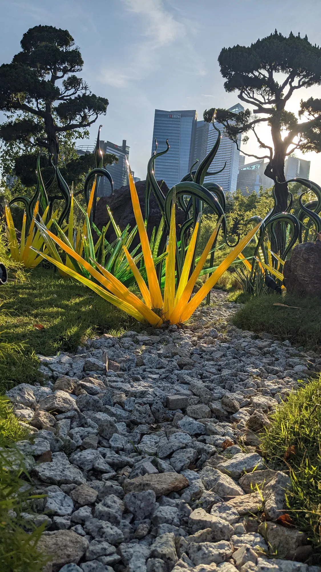 A spiky yellow glass sculpture in a garden with trees and towers behind
